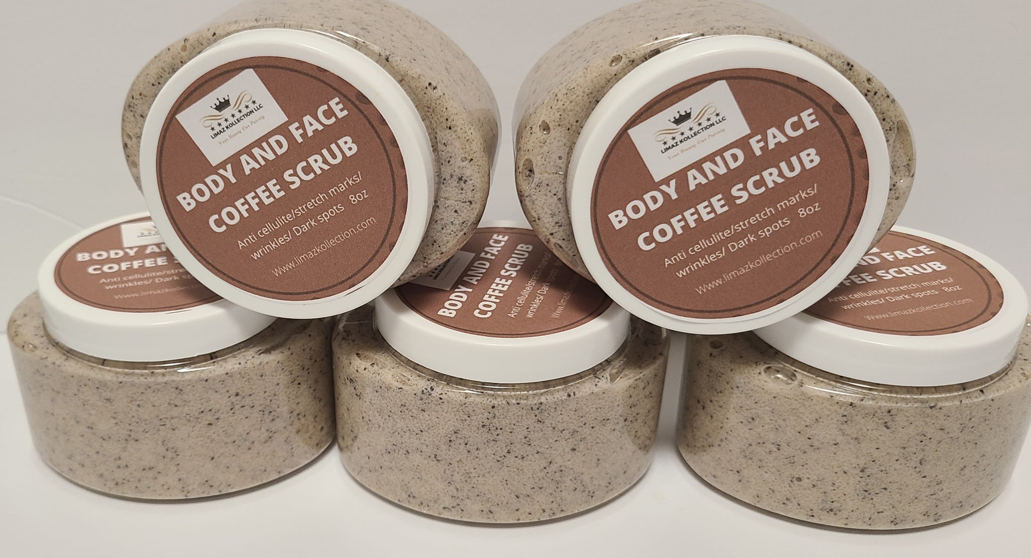 The image showcases a collection of four jars of “Body and Face Coffee Scrub” by “Limax Kollection LLC”. The jars are cylindrical with dark brown lids and clear bases, allowing the scrub’s texture to be visible. The scrub itself appears to be a granular mixture of brown and beige tones, suggesting coffee grounds combined with other ingredients. Each jar is adorned with a rectangular label featuring a dark red background and white text. The label prominently displays the product name, “BODY AND FACE COFFEE SCRUB”, in a bold, stylized font. Below the name, it lists the scrub’s benefits: “Anti cellulite/stretch marks/ wrinkles/ Dark spots 8oz”. The Limax Kollection LLC logo, consisting of a crown and stylized text, is positioned at the top of the label. The website address, “www.limaxkollection.com”, is printed at the bottom. The jars are arranged in a slightly staggered formation on a neutral, light-colored surface. Three jars are fully visible, while a portion of the fourth is seen in the foreground. The lighting is bright and even, highlighting the texture of the scrub and the details of the labels. The overall presentation suggests a focus on natural skincare and a homemade, artisanal quality. The arrangement implies a product line or a set for sale.